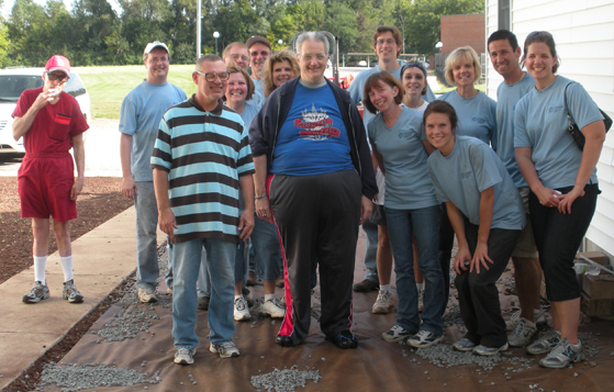 A group of people standing in front of a white house. There are around 20 people in the group all wearing blue t-shirts and smiling at the camera. In the center of the group is an older man wearing a blue shirt with a red and black striped shirt and black pants. He is standing with his hands in his pockets and appears to be posing for a group photo. To his left there is a man in a red jumpsuit and a baseball cap who is holding a baseball bat. Behind him there are trees and a car parked on the side of the house. The ground is covered in gravel and there are a few pieces of debris scattered around.