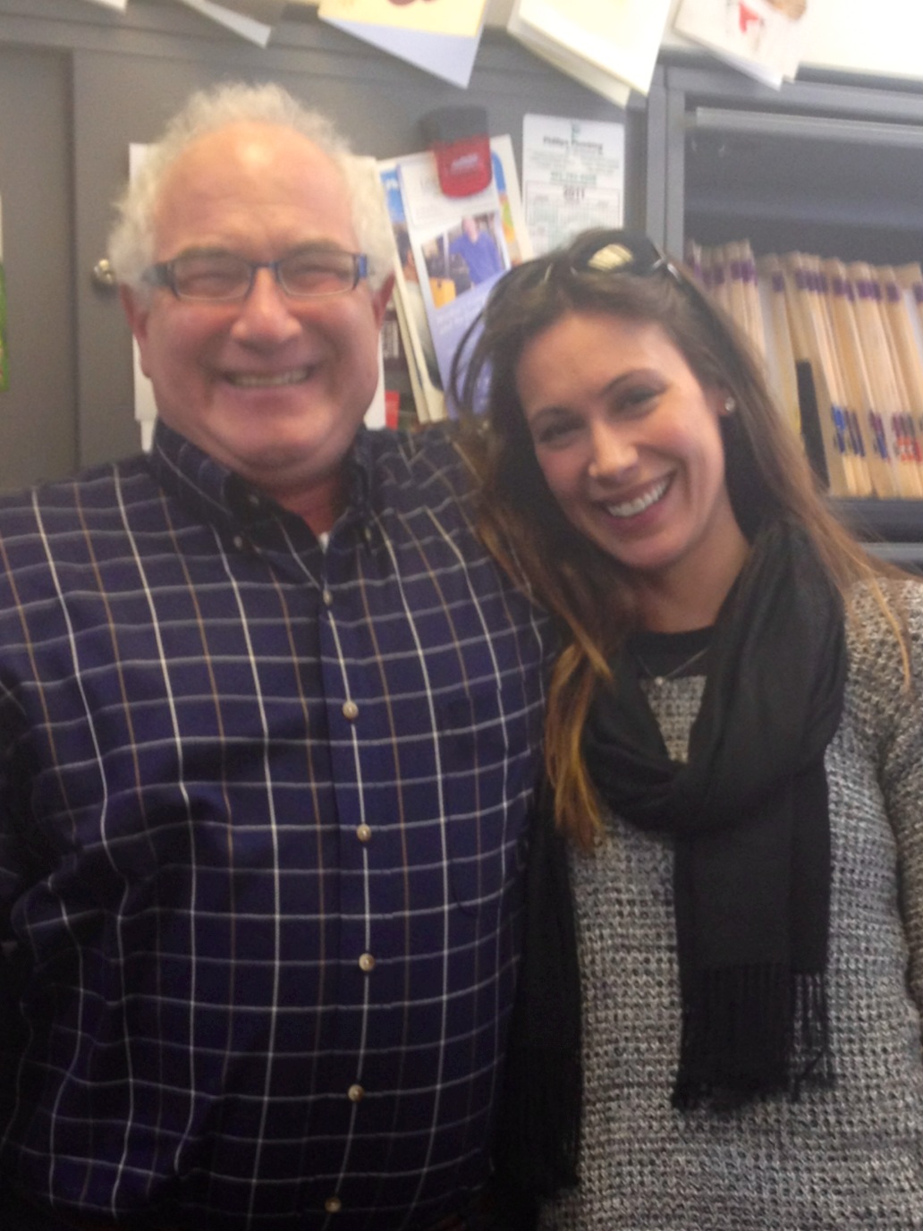 A man and a woman standing side by side and smiling at the camera. The man is on the left side of the image and the woman on the right side. They are standing in front of a bookshelf filled with books and papers. The woman is wearing a black scarf and a gray sweater and she has long brown hair. Both of them are wearing glasses and appear to be happy and relaxed. The background is blurred but it appears to be an office or a library.