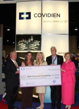 A group of five people standing in front of a large banner that reads "Covidien Positive Results for Life". The banner also has an image of a bottle of pills and the logo of the company. The group is holding a large check for $15000. The check is being presented to the AHA Education Foundation. The people in the group are smiling and appear to be proud of the donation. The background is a blue carpeted floor and there are other people visible in the background.