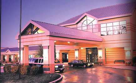 The exterior of a hotel at night. The hotel is a two-story building with a modern design featuring a curved roof and large windows on the second floor. The building is painted in a combination of orange and beige colors with a red stripe running along the side. The entrance of the hotel is covered by a covered walkway and there are two cars parked in front of the entrance. The sky is dark indicating that it is either dusk or early morning. There is a street lamp on the left side of the image and a few trees and bushes in the foreground.