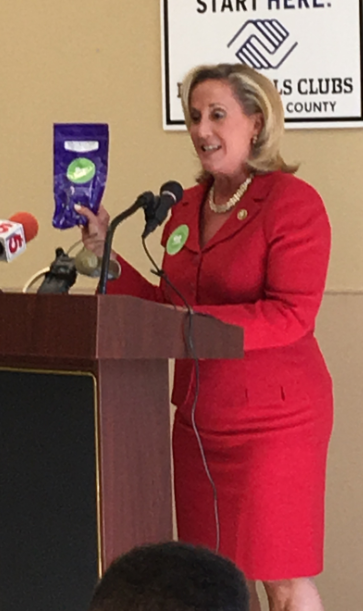 A woman standing behind a podium and speaking into a microphone. She is wearing a red suit and a pearl necklace. She has blonde hair and is holding a purple packet of chips in her hand. Behind her there is a sign that reads "Start Here! Hills Clubs County" and a banner with the Hills Clubs logo. There are several microphones in front of her indicating that she is giving a speech or presentation. The background appears to be a room with a beige wall and a window.