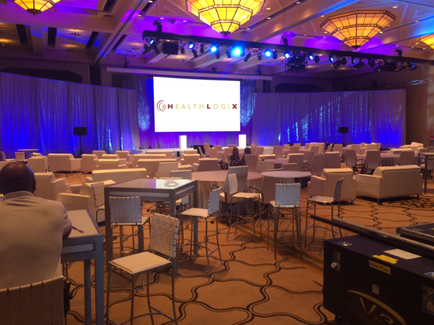 A large conference room with a high ceiling and multiple chandeliers hanging from it. The room is set up with white chairs and tables arranged in rows facing a stage. On the stage there is a large screen with the word "HEALTHLOGIX" written in red and white letters. The stage is decorated with blue curtains and there are speakers on either side of the screen. A man is sitting at a desk in front of the stage and appears to be giving a presentation. The floor is covered with a patterned carpet.