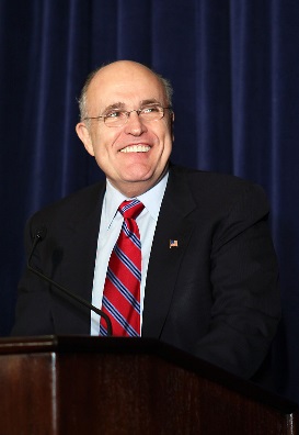 A middle-aged man standing behind a wooden podium and speaking into a microphone. He is wearing a dark suit and a red and blue striped tie. He has a small American flag pin on his lapel and is smiling widely. The background is a blue curtain. The man appears to be giving a speech or presentation.