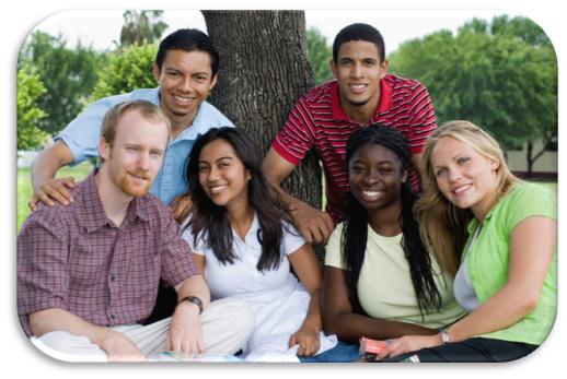 A group of six young people two men and three women sitting together under a tree in a park. They are all smiling and appear to be happy and relaxed. The man on the left is wearing a plaid shirt and has a beard while the man in the center is leaning against the tree with his arms around the woman on the right. The woman in the middle is sitting on the ground with her legs crossed and is holding a book or notebook. The other three people are standing behind her with one man wearing a red and white striped shirt and the other two wearing casual clothes. The background is filled with trees and greenery suggesting that the photo was taken in a natural setting.