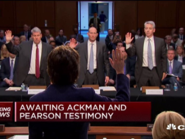 A group of four men standing in a courtroom with their hands raised in the air. They are all wearing suits and appear to be in the middle of a hearing. The man on the left is wearing a red tie and the man in the center is wearing glasses. The other three men are wearing suits. The background shows a large crowd of people sitting in the courtroom. At the bottom of the image there is a news headline that reads "Awaiting Ackman and Pearson testimony."