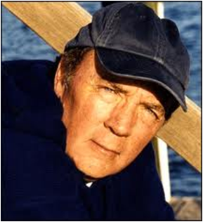 A close-up portrait of a middle-aged man wearing a black baseball cap. He appears to be in his late 60s or early 70s with short dark hair and a serious expression on his face. He is leaning against a wooden railing with the ocean visible in the background. The man is wearing a dark blue jacket and his eyes are looking off to the side. The overall mood of the image is somber and contemplative.