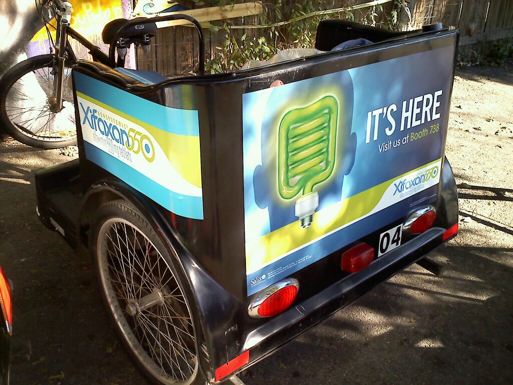 A small black tricycle with a blue and yellow advertisement on the back. The advertisement features a green microphone with the words "It's Here" written above it. The tricycle is parked on a street with a wooden fence and a bicycle in the background. The license plate reads "04".