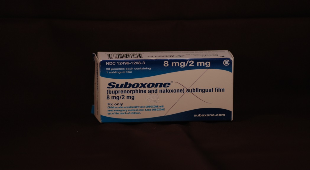 A photograph of a white box with blue text on it. The box is labeled "Suboxone" and has a barcode on the top left corner. The text on the box reads "8 mg/2 mg" in bold letters. Below the text there is a description of the product which states that it is a sublingual film. The label also mentions that the product is "buprenorphine and naloxone (sublingual) film". The box appears to be sitting on a black surface.