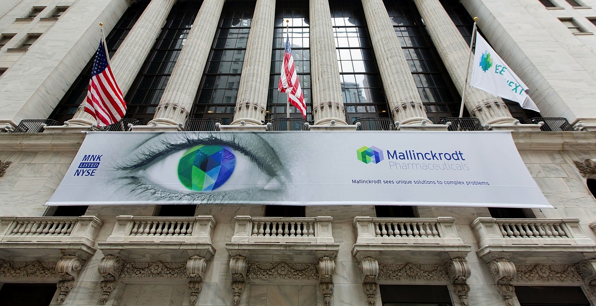 The exterior of the New York Stock Exchange building in New York City. The building has a large banner hanging from the side of the building with the logo of Mallinckrodt Pharmaceuticals a multinational pharmaceutical company on it. The banner also has an image of an eye with a rainbow-colored iris in the center. Above the eye there are three American flags flying in the air. The American flag on the left is the American flag the one on the right is the United States flag and the one in the middle is the logo for the company. The sky is blue and the building is made of stone with intricate carvings and columns.