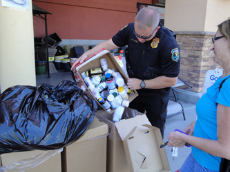 A police officer in a black uniform with a badge on his chest standing next to a woman in a blue shirt. The officer is holding a cardboard box filled with various items including bottles cans and other household items. The woman is standing in front of a pile of cardboard boxes and a black garbage bag. They appear to be sorting through the items in the box. In the background there is a building with a sign that reads "GOODS".