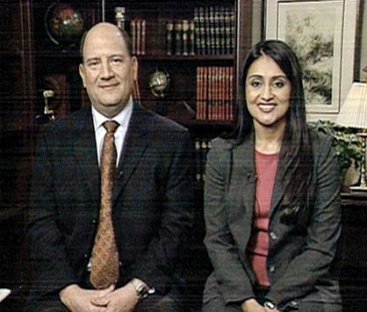 A man and a woman sitting side by side in a room with a bookshelf in the background. The man is on the left side of the image and the woman on the right side. They are both dressed in formal attire with the man wearing a suit and tie and the women wearing blazers. They both have serious expressions on their faces and are looking directly at the camera. The room appears to be a formal setting with a desk a lamp and a framed picture on the wall.