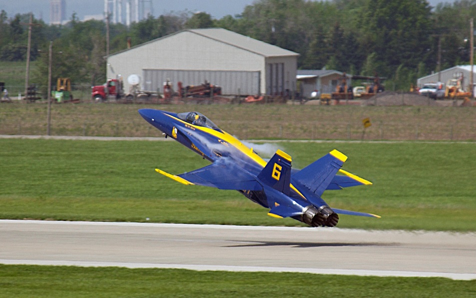 A blue and yellow fighter jet taking off from an airport runway. The jet is in the center of the image with its wings spread wide and its nose pointed upwards. The tail of the jet is painted in the colors of the United States Navy with the number 6 on the side. In the background there is a large hangar and a construction site with various vehicles and equipment. The sky is overcast and there are trees and buildings visible in the distance.