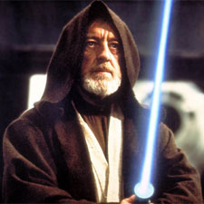 A still from the movie Star Wars: Episode IV - A New Hope. It shows the main character Obi-Wan Kenobi holding a blue lightsaber in his right hand. He is wearing a black robe with a hood and has a white beard and mustache. Kenobi is looking directly at the camera with a serious expression on his face. The background is blurred but it appears to be a scene from the Star Wars universe.