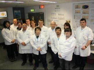 A group of people standing in a laboratory. They are all wearing white lab coats and are posing for a group photo. There are nine people in the photo nine men and one woman all of whom appear to be of different ages and ethnicities. The group is standing in front of a whiteboard with various scientific notes and diagrams pinned to it. There is a sign above the group that reads "Exit" and a door in the background. The overall mood of the photo is serious and professional.