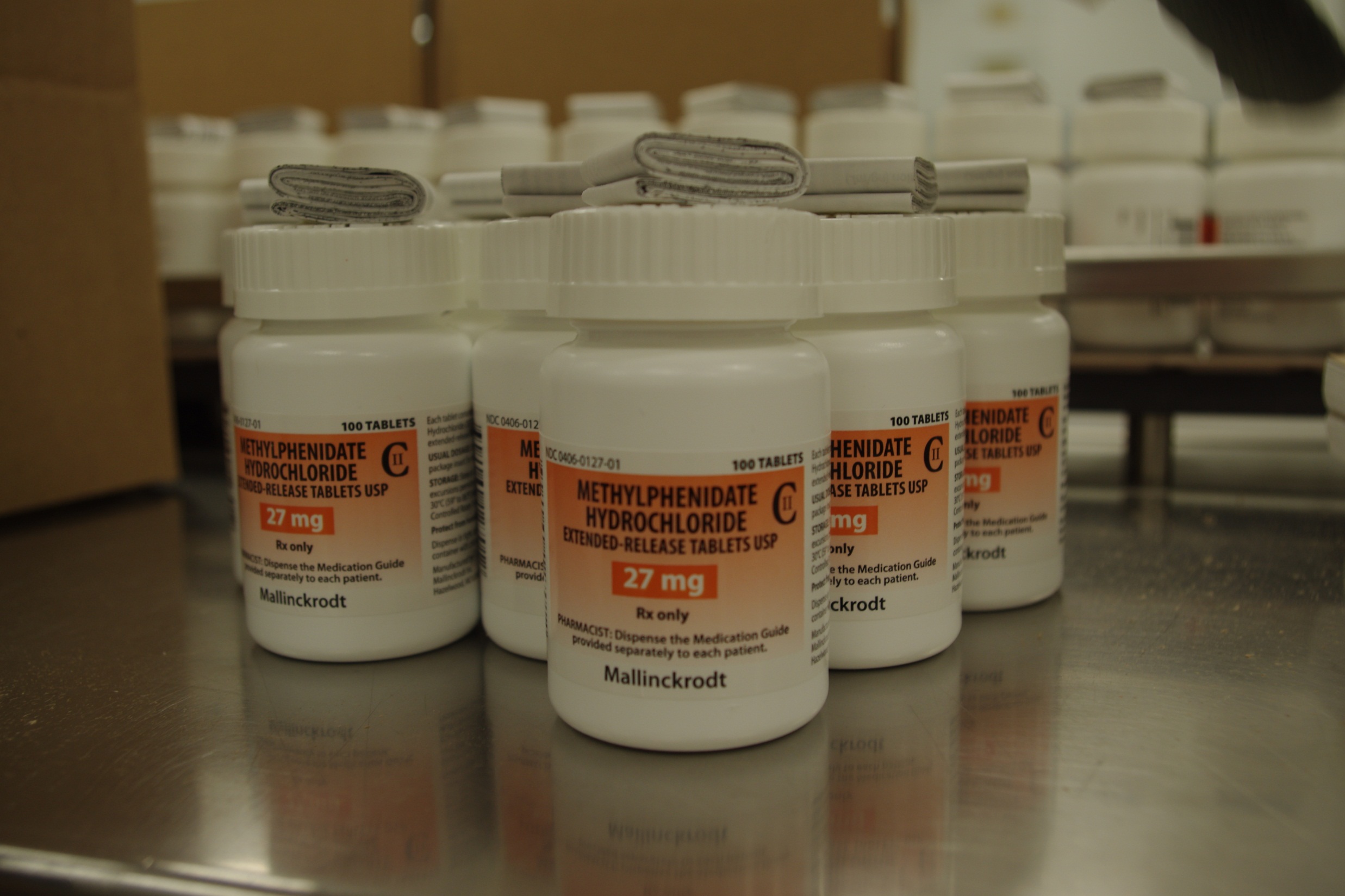 A row of white plastic bottles with orange labels on them. The bottles are arranged in a neat row on a metal countertop. The labels on the bottles read "Methylphenidate Hydrochloride 27 mg" and "Mallinckrodt". The bottles appear to be new and unused. In the background there are more bottles of the same product and a cardboard box. The image appears to be taken in a laboratory or a medical facility.