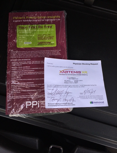 A red packet of Patient Prescription Insights with a green label that reads "Your first Rx free". Next to the packet there is a white piece of paper with the text "XARTEMIS XR" and "Physician Stocking Request" written on it. The packet appears to be sealed and is placed on a black surface. The background is blurred but it seems to be the interior of a car.