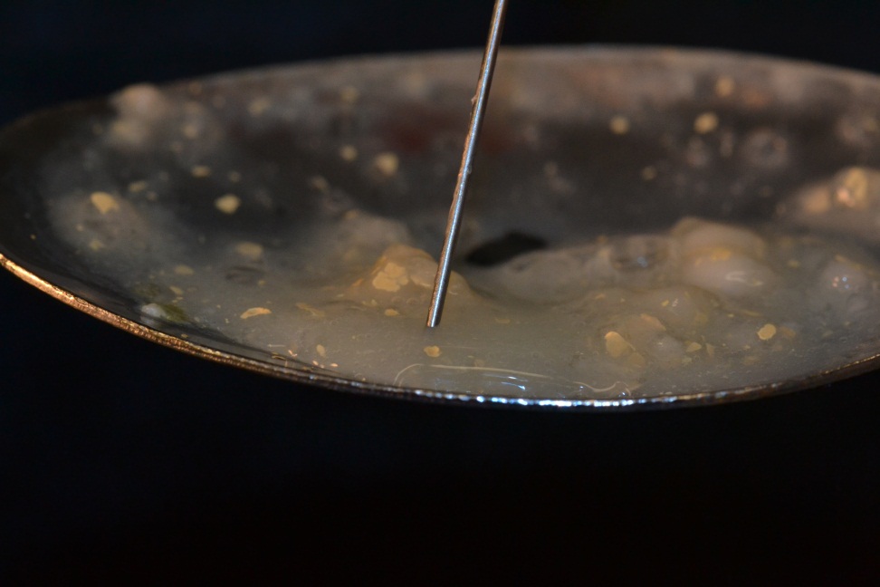 A close-up of a frying pan with a small amount of food in it. The pan appears to be made of stainless steel and has a shiny surface. The food in the pan is a dark brown color and there are small pieces of meat and vegetables visible in the mixture. A metal spoon is resting on the edge of the pan and it seems like someone is about to stir the food. The background is dark and out of focus making the pan the focal point of the image.