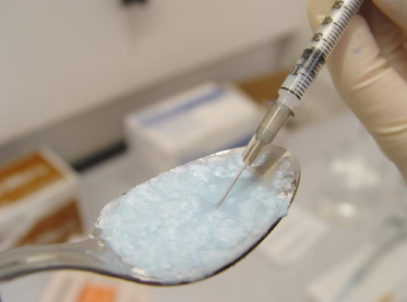 A spoon filled with a light blue substance likely an opioid or another drug and a hand holding a syringe. The syringe drawing up the substance.  The hand is wearing a white safety glove. The background is blurred but it appears to be  laboratory setting.