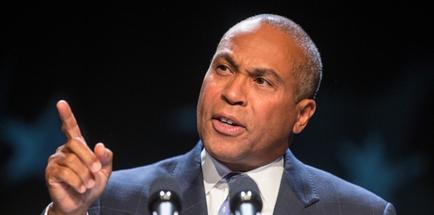A middle-aged African-American man speaking at a podium with microphones in front of him. He is wearing a suit and tie and appears to be giving a speech or presentation. The background is dark and blurred suggesting that the focus is on the man and his speech. He has a serious expression on his face and is pointing upwards with his right hand as if he is making a point or making a statement.