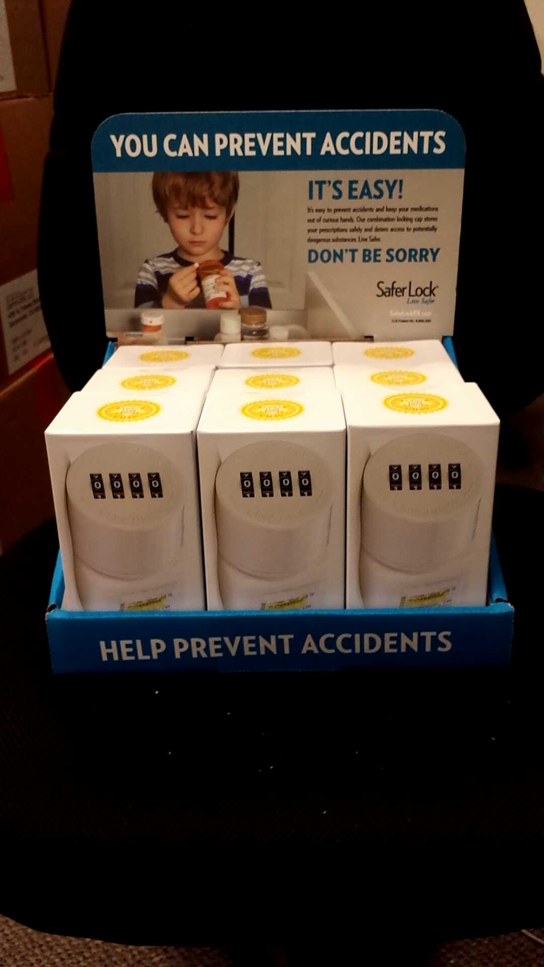 A blue cardboard box with a label that reads "You can prevent accidents. It's easy! Don't be sorry. Safer Lock." Inside the box there are four white boxes with yellow labels. The boxes are arranged in a pyramid-like shape and appear to be made of plastic. The label on the top of the box has an image of a young boy holding a cup of coffee and looking at the camera. The background is blurred but it appears to be an indoor setting.