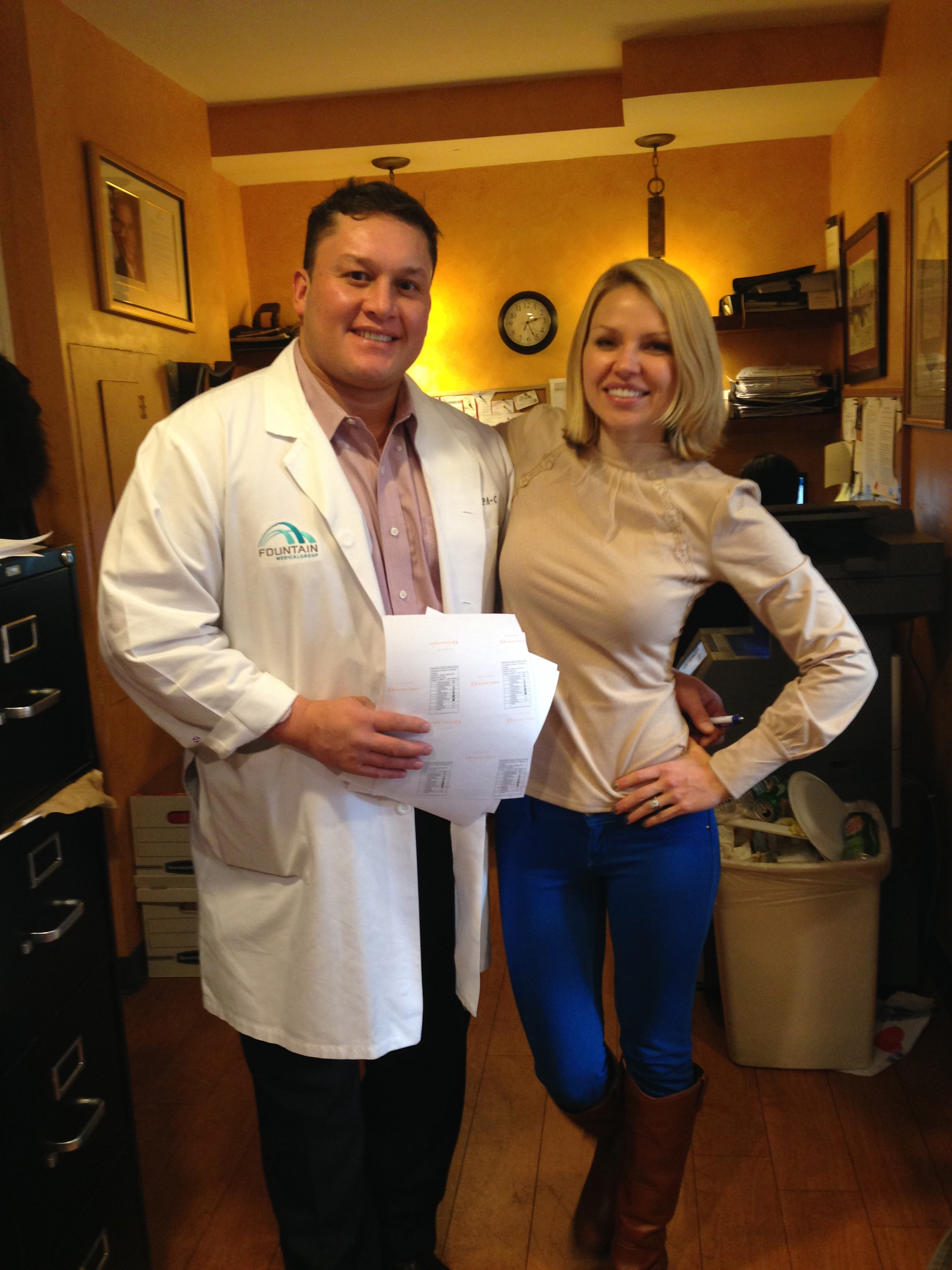 A man and a woman standing in a room with orange walls and wooden flooring. The man is wearing a white lab coat with a blue logo on the left side of his chest and is holding a piece of paper in his left hand. He is smiling at the camera and the woman is standing next to him with her right hand on his hip. They are both dressed in formal attire and appear to be posing for a photo. In the background there is a desk with a computer and a clock on the wall.