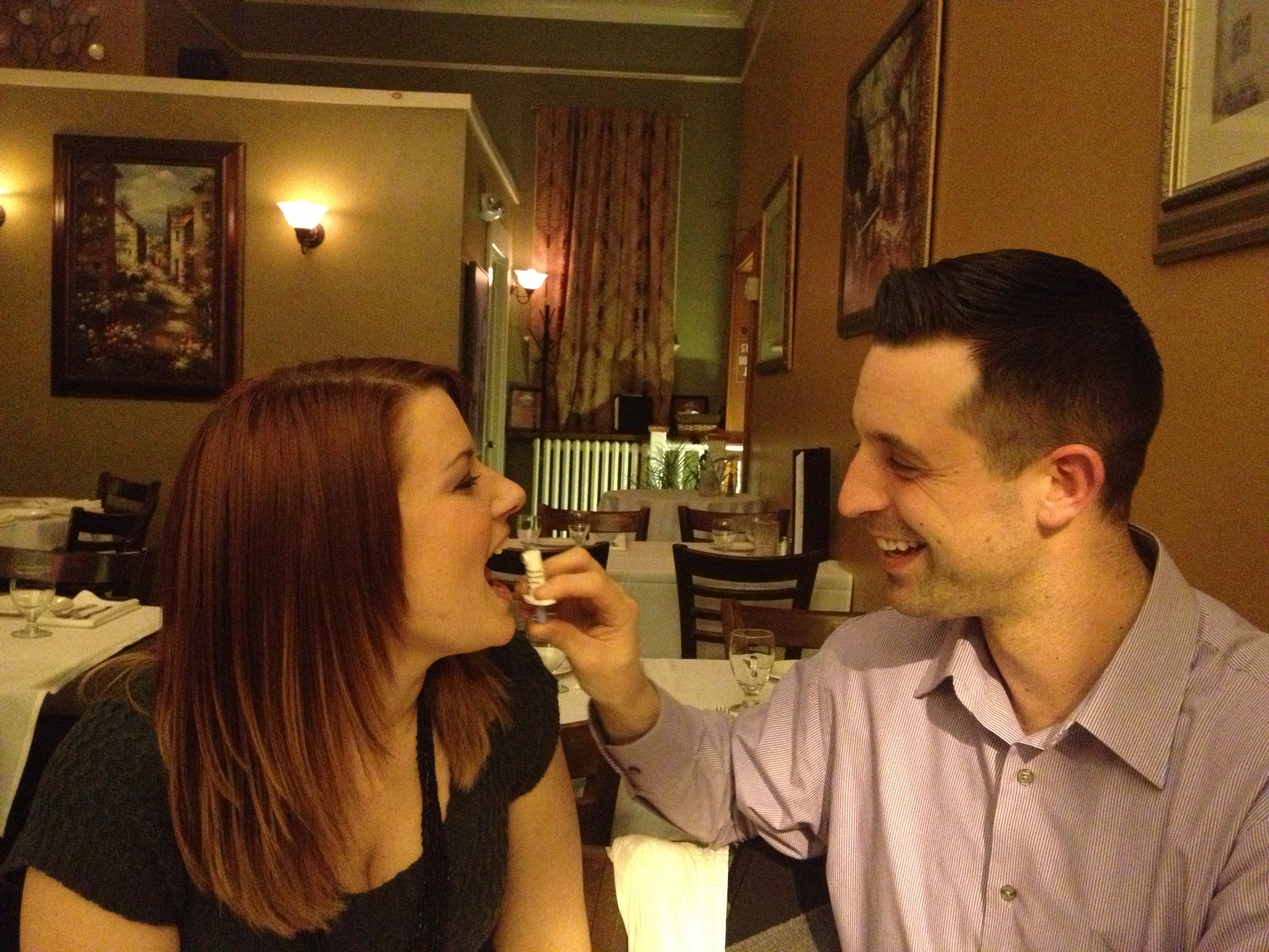 A young couple sitting at a table in a restaurant. The woman is on the left side of the image with long red hair and is wearing a black top. She is holding a small white object in her hand and appears to be taking a bite out of it. The man on the right side is sitting next to her with a smile on his face. They are both looking at each other and seem to be enjoying each other's company. The restaurant has wooden tables and chairs and there are several framed pictures hanging on the walls. The overall atmosphere of the restaurant is cozy and intimate.