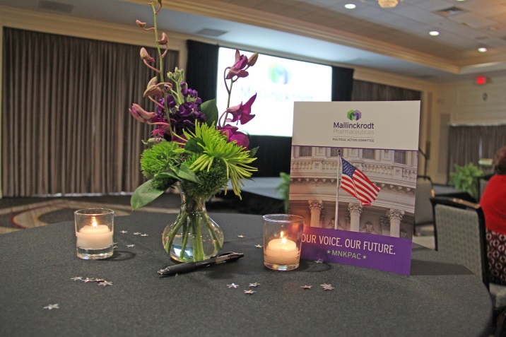 A table with a vase of purple and green flowers and two small candles on it. The table is covered with a black tablecloth and there are small white stars scattered around the table. In the background there is a large screen and a banner with the text "Our voice our future" and an image of an American flag. The room appears to be a conference room with a high ceiling and a chandelier hanging from the ceiling.