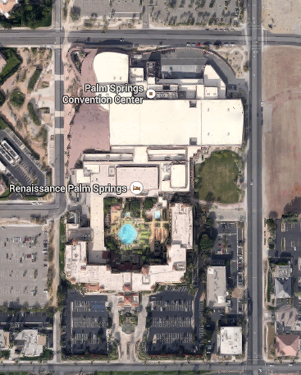 An aerial view of the Renaissance Palm Springs Convention Center. The center is located in the center of the image and is surrounded by several buildings and roads. The building on the left is a large white building with a curved roof while the one on the right is a smaller white building. There is a blue swimming pool in the middle of the center and several smaller buildings surrounding it. The image is taken from a bird's eye view looking down on the convention center and the surrounding area.