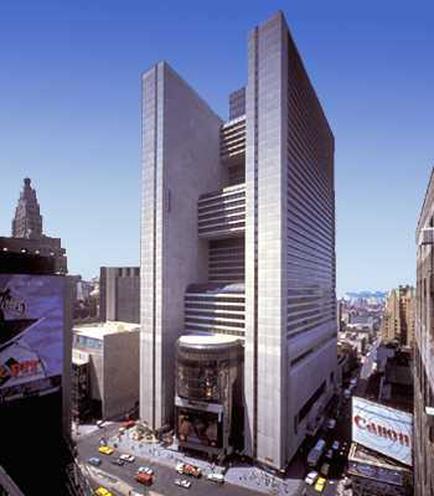 An aerial view of a tall modern building in a busy city. The building appears to be a high-rise with multiple floors and a unique architectural design. It is located in the center of the image surrounded by other buildings and skyscrapers. The sky is clear and blue and there are a few cars and trucks on the street below. On the left side of the building there is a large billboard with the Canon logo and on the right side there are several other buildings. The image appears to have been taken from a high vantage point looking down on the city below.