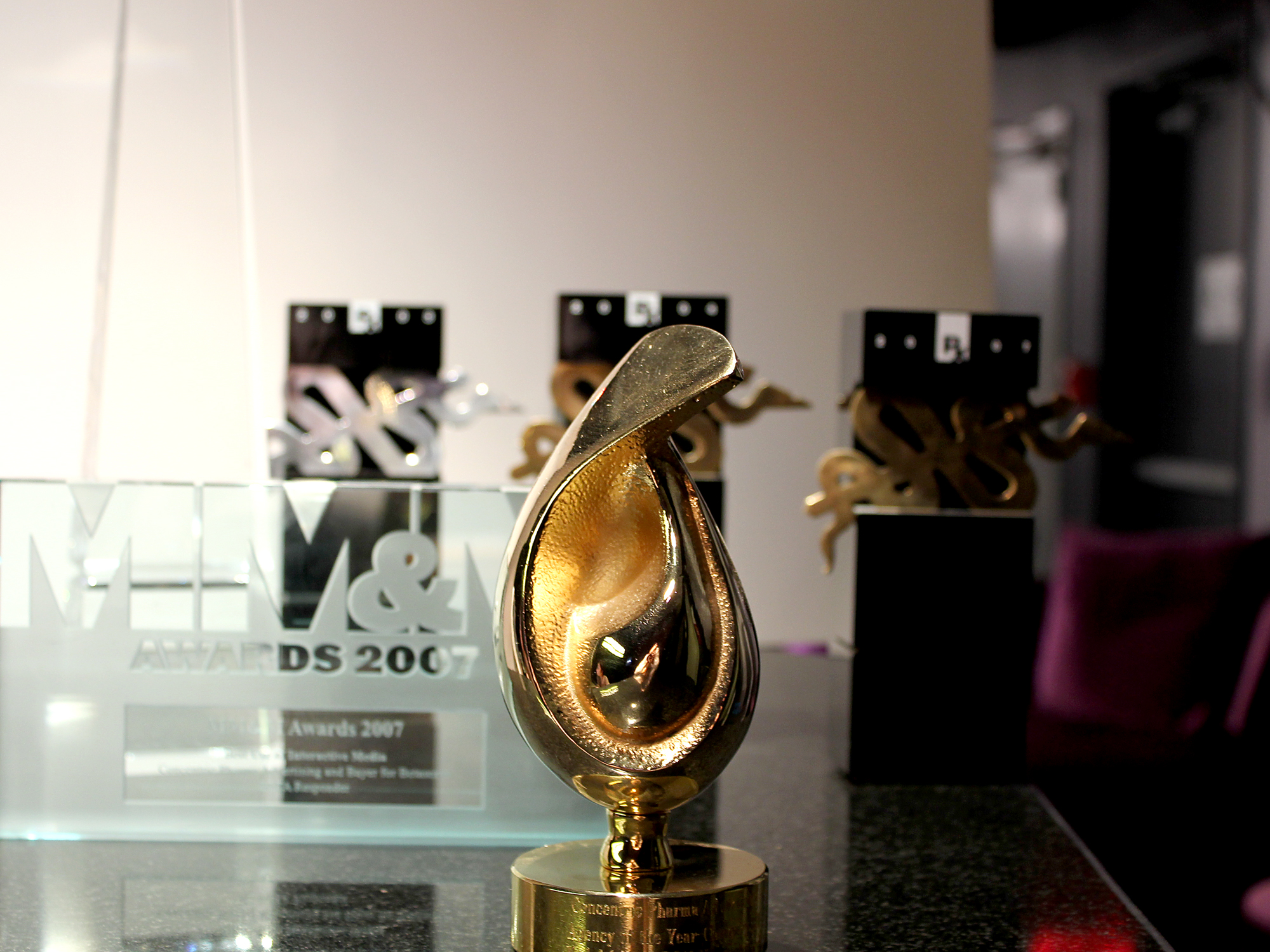 A gold-colored trophy on a black countertop. The trophy is in the shape of a teardrop with a curved top and a pointed bottom. It is placed on a clear glass base with the words "M&M Awards 2007" engraved on it. In the background there are three other trophies one of which is a black and gold design. The background is blurred but it appears to be a room with a white wall and a pink chair.