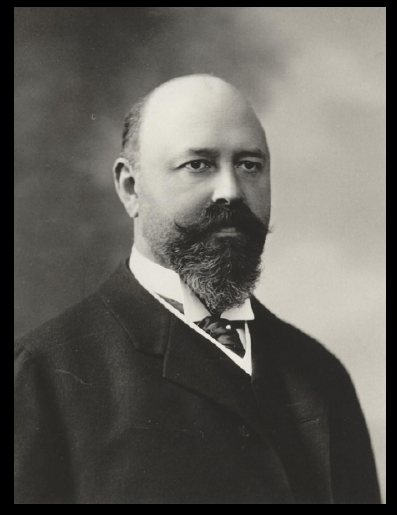 A black and white portrait of a man. He is facing the camera and is wearing a suit and tie. The man has a full beard and mustache and is bald. He has a serious expression on his face and is looking directly at the camera. The background is blurred so the focus is on the man's face. The image appears to be a studio portrait.