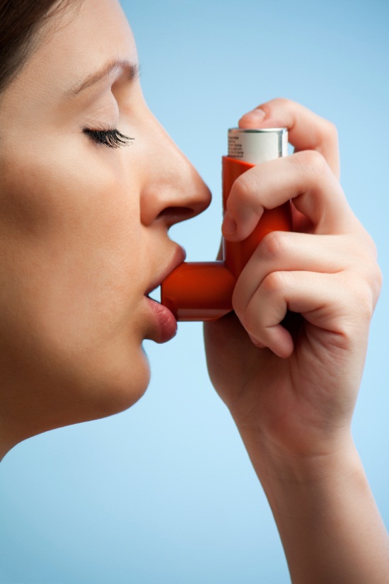 A close-up of a woman's face with her eyes closed and her mouth slightly open as she uses an asthma inhaler. The inhaler is orange in color and is held in XX her right hand. The background is a solid blue color. The woman appears to be in her late twenties or early thirties and has dark hair. She is holding the inhaler with both hands and is using it to inhale the air.