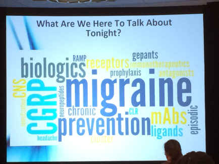 A large screen with a presentation slide on it. The slide has a blue background with white text that reads "What Are We Here To Talk About Tonight?" The text is arranged in a circular pattern and is in different sizes and colors. The title of the slide is "Migraine Prevention" in large bold letters at the top. Below the title there is a list of topics related to migraine prevention such as "biologic" "ramp receptors" "tenants" "prophylaxis" "antagonists" "chronic" "clr" "mass" "headache" and "cluster". The slide appears to be from a conference or presentation as there are people visible in the background.