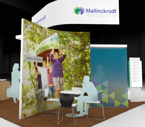 A trade show booth with a large banner on the left side of the booth. The banner has a photo of a family of three - a man a woman and two children - standing in front of a green background with trees and foliage. The man is holding up a sign that reads "Mallinckrodt Pharmaceuticals" and the children are smiling and waving to the camera. On the right side there is a blue banner with a geometric design and a white table and chairs. The booth is set up in a room with a wooden floor and a black wall in the background.