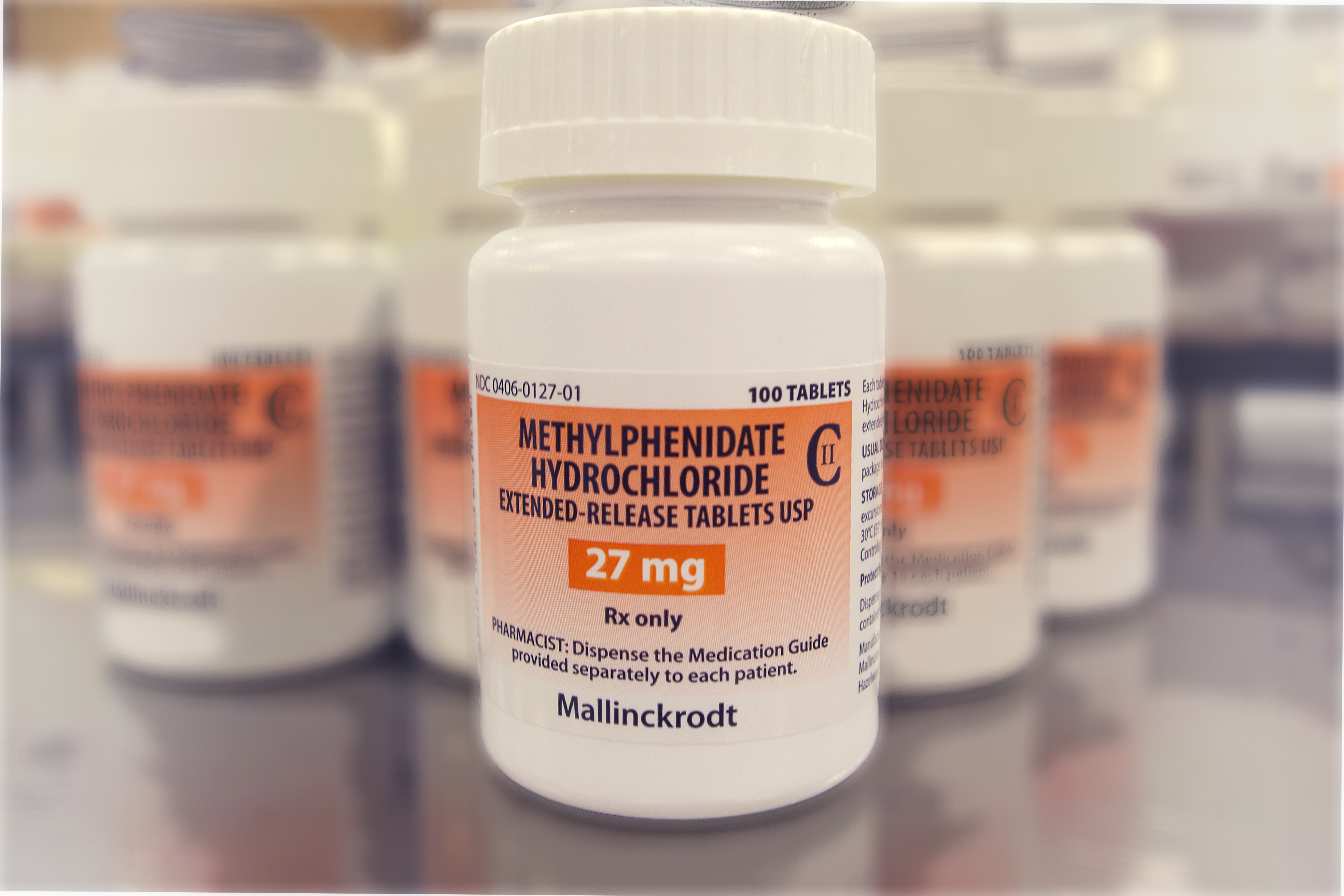 A white medicine bottle with a white cap. The label on the bottle reads "Methylphenidate Hydrochloride Extended-Release Tablets USP 27 mg". The label also mentions that the bottle contains Rx only. In the background there are several other white medicine bottles with orange labels. The bottles are arranged in a row on a shelf.