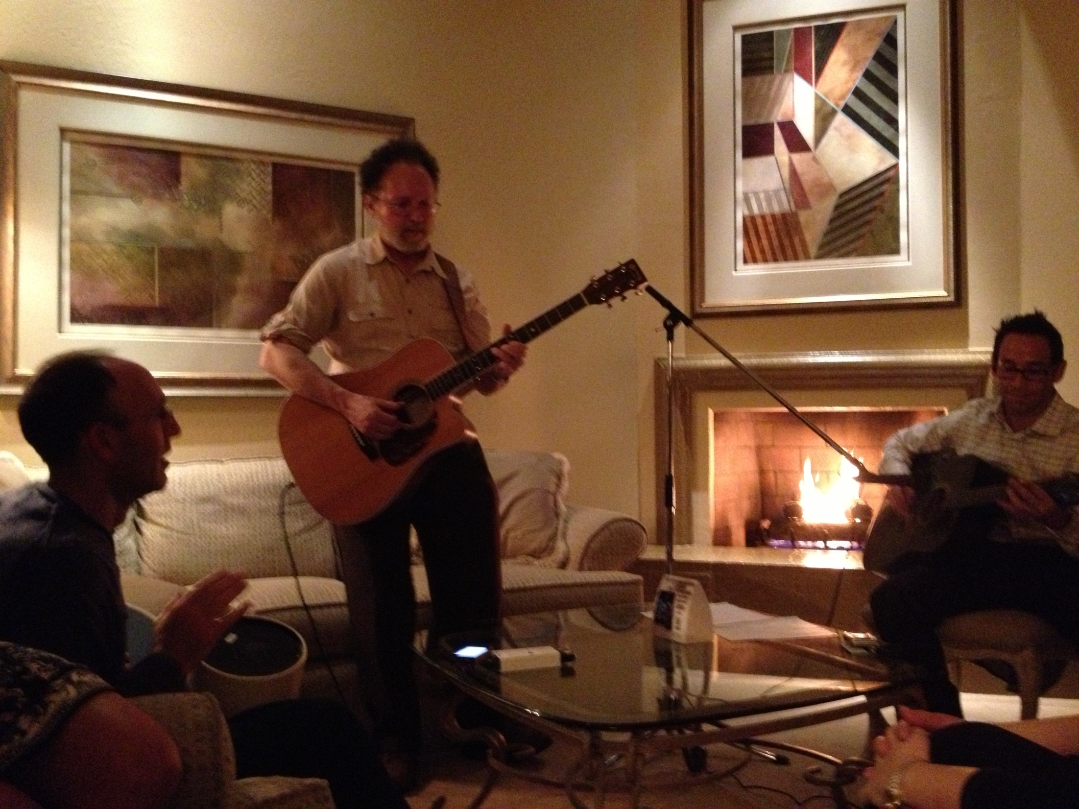 A group of three men in a living room. The man in the center is playing an acoustic guitar and singing into a microphone. He is wearing a beige shirt and glasses and appears to be in the middle of a performance. To his left there is a man sitting on a couch and playing a guitar and to his right there are two other men sitting on chairs and playing guitars. The room is decorated with a fireplace and a painting hanging on the wall. 