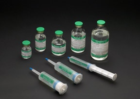 A group of six small glass vials with green caps and white labels. The vials are arranged in a scattered manner on a black background. There are six of the vials in total with the largest one in the center and three smaller ones on either side. The first vial on the left has a green cap the second one has a white cap and the third one is a clear plastic vial with a blue cap. All the vials appear to be filled with a clear liquid.