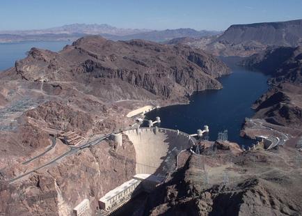 A large dam located in the middle of a large body of water. The dam is surrounded by rocky mountains on both sides and the water is a deep blue color. The sky is clear and blue and the mountains in the background are a mix of brown and gray. On the left side of the image there is a winding road that leads to the dam and on the right side there are several buildings and structures scattered around the area. The water is calm and still reflecting the sky above. The image appears to be taken from a high vantage point looking down on the dam and the surrounding landscape.
