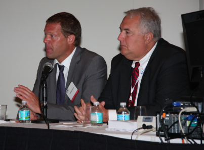 Two men sitting at a table with a microphone in front of them. They are both wearing suits and appear to be engaged in a discussion. The man on the left is speaking into the microphone and has a name tag around his neck. He is gesturing with his hands as he speaks while the other man is listening attentively. On the table there are water bottles and a computer monitor. The background is a plain white wall.