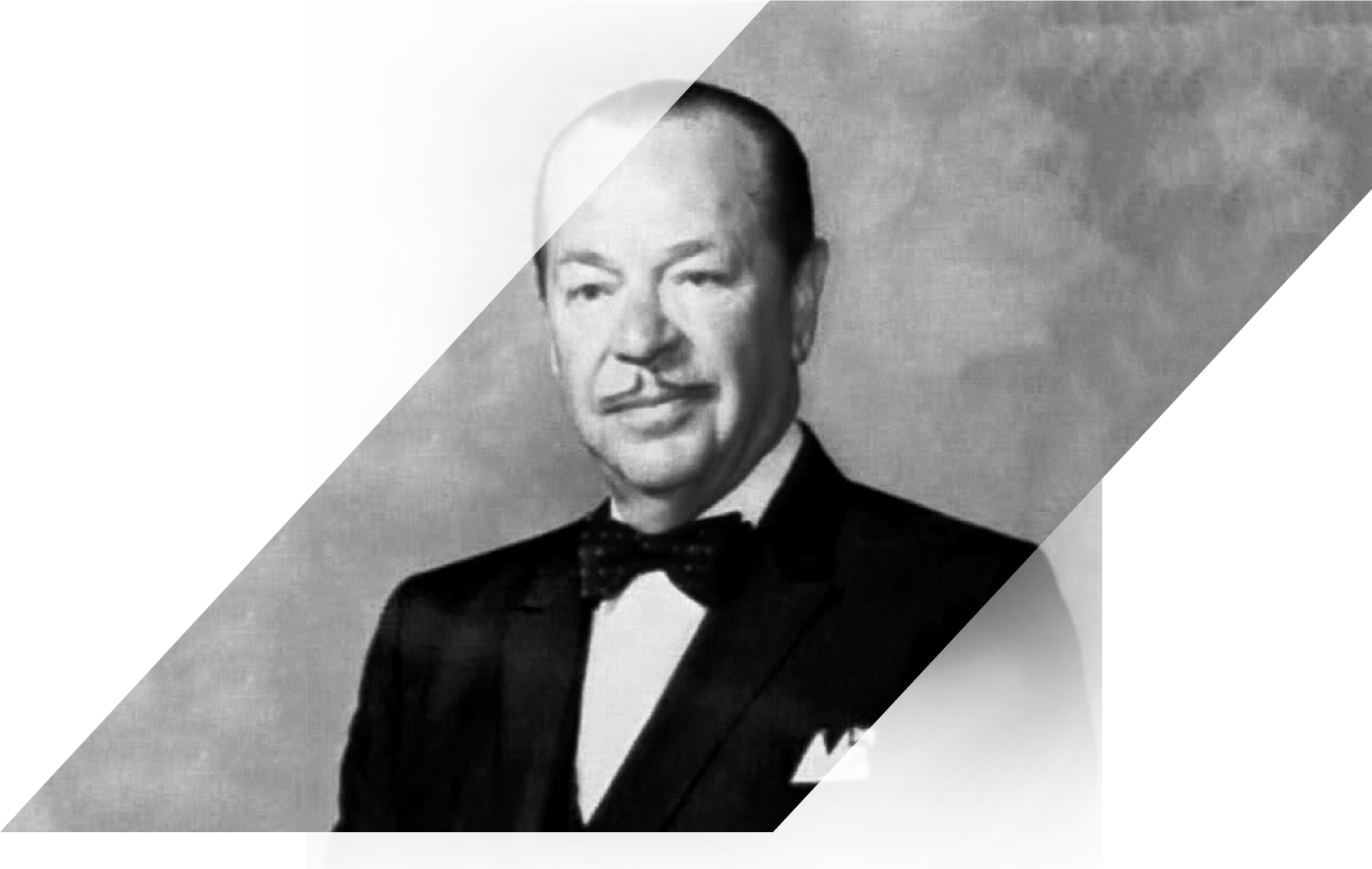 A black and white portrait of a man in formal attire. He is wearing a tuxedo with a white shirt and a black bow tie. He has a mustache and is looking directly at the camera with a slight smile on his face. The background is a plain grey color. The man appears to be in his late 60s or early 70s.