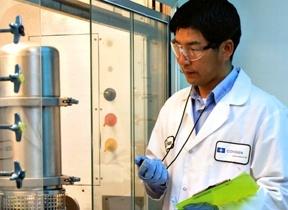 A man in a laboratory setting. He is wearing a white lab coat a stethoscope around his neck and blue gloves. He appears to be a scientist or a researcher as he is holding a green folder in his left hand and is looking down at something in his right hand. In the background there is a large stainless steel tank with pipes and valves and a control panel with various buttons and switches. The man seems to be focused on his work.