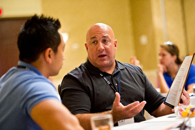 A man sitting at a table and talking to another man. The man in the foreground is bald and wearing a black polo shirt. He is holding a menu in his hand and appears to be explaining something to the other man who is wearing a blue t-shirt. There are other people sitting at the table in the background but they are not visible. The table is covered with a white tablecloth and there are glasses of water on it. The room appears to have a beige wall and a door.