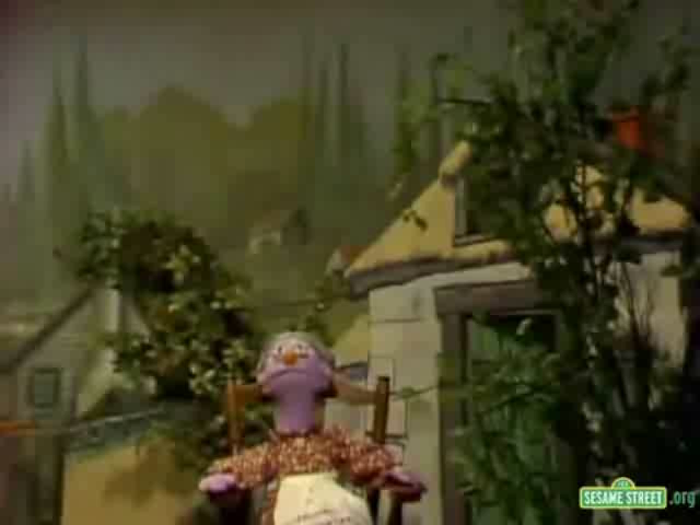 A still from the Sesame Street animated series. It shows a scene from the show where the main character Cookie Monster is sitting on a wooden chair in front of a house. Cookie Monster is wearing a pink dress with a white apron and has a big smile on his face. He is holding a bouquet of flowers in his hands. The house is a two-story building with a yellow roof and a chimney. There are trees and bushes in the background and a mountain range can be seen in the distance. The overall mood of the image is cheerful and playful.