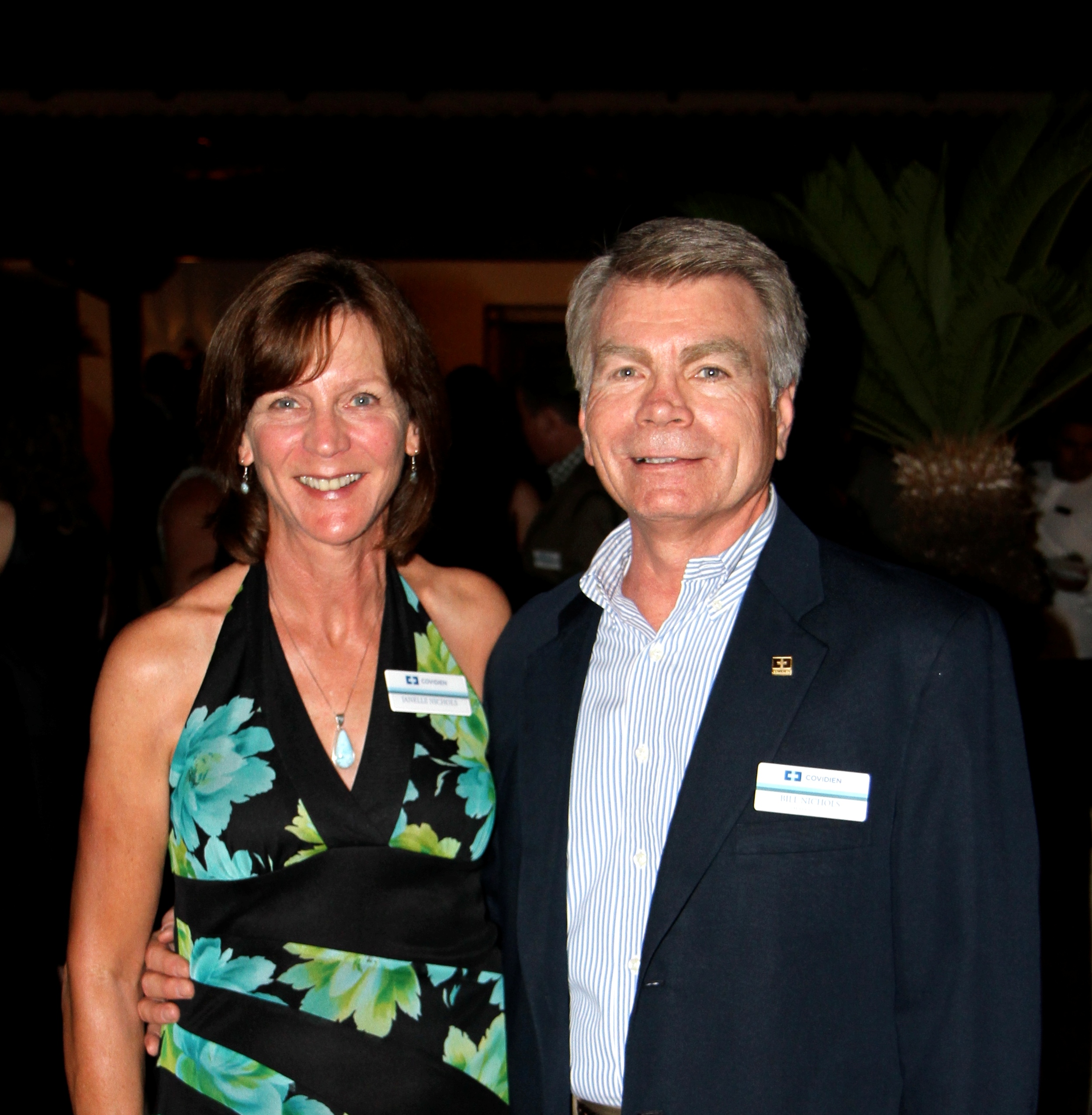 A man and a woman standing side by side and smiling at the camera. The woman is on the left side of the image and the man on the right side. They are both dressed in formal attire with the woman wearing a black and green floral dress and a necklace. The man is wearing a blue blazer with a white shirt and a name tag on his lapel. They appear to be at an event as there are other people in the background.