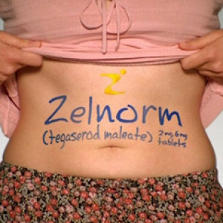 A woman's stomach with the word "Zelnorm" written on it in blue ink. The word is written in a cursive font with a yellow letter "Z" in the center. Below the word there is a smaller text that reads "Tegasorod maleate" and "2mg tablets". The woman is wearing a pink top and a floral skirt. She is holding her stomach with both hands and appears to be in pain or discomfort. The background is plain white.