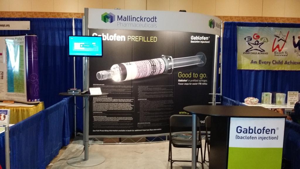 A trade show booth with a large banner that reads "Mallinckrodt Pharmaceuticals" and "Gablofen Prefilled". The banner also has an image of a syringe with the text "Good to go" above it. The booth is set up in a room with blue curtains and there is a table in front of it with a sign that says "Gablofen" on it. There is also a TV screen on the left side of the booth and a banner on the right side.