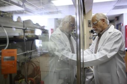 Two elderly men in a laboratory setting. They are both wearing white lab coats and glasses and appear to be working together on a project. The man on the left is standing in front of a large glass window and he is looking through it intently at something on the right side of the image. Behind him there are various scientific equipment and equipment including pipes tubes and other scientific equipment. The room is dimly lit and there is a red fire extinguisher visible in the background. The men seem to be focused on their work and are engaged in a conversation.