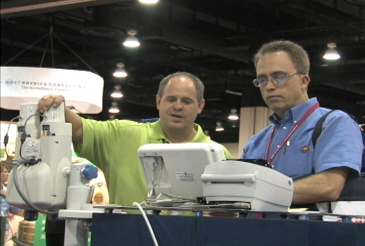 Two men standing in front of a table with a machine on it. The man on the left is wearing a green polo shirt and is holding the machine with both hands. He appears to be explaining something to the other man who is also wearing a blue shirt and glasses. The machine is white and has a control panel on the front. The table is covered with a blue tablecloth and there are other machines and equipment visible in the background. There is a banner hanging from the ceiling with the words "Westphalia Construction" written on it indicating that the image was taken at a trade show or exhibition.