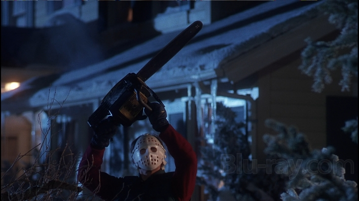 A person wearing a Jason Voorhees mask and holding a chainsaw above their head. The person is standing in front of a house with a porch covered in snow. The house is lit up with a street lamp and there are trees in the background. The image appears to be a still from a horror movie or TV show.