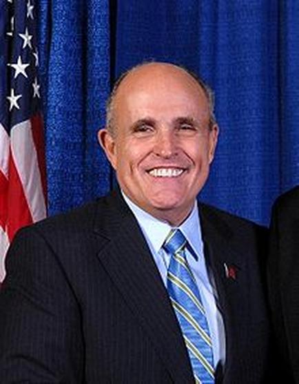 A portrait of a middle-aged man in a suit and tie. He is standing in front of an American flag and a blue curtain. The man is smiling widely and appears to be in his late 60s or early 70s. He has a bald head and is wearing a dark suit with a blue shirt and a yellow and blue striped tie. There is a small red pin on his lapel. The image appears to have been taken in a formal setting possibly a government or government office.